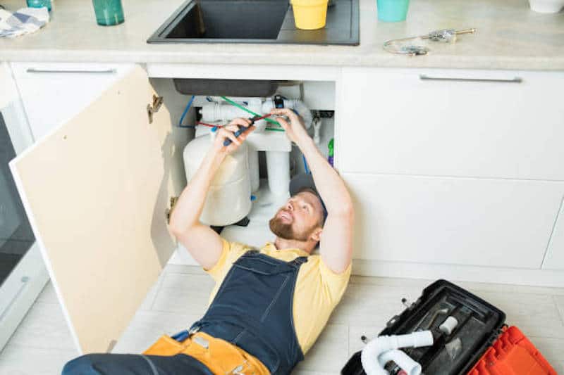 Positive bearded plumber in uniform lying on flor and cutting cables while fixing water pipe in kitchen