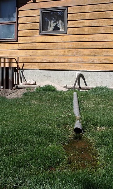 a tube comes out of a wood sided house - emptying sludge from teh sump pump onto green grass