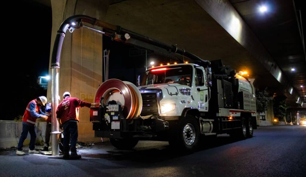 Two workers operate a street cleaning truck under a bridge at night.